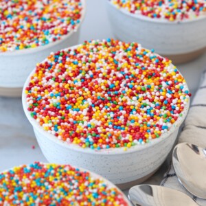 A white bowl filled with a dot cake topped with a dense layer of colorful round sprinkles; more bowls and spoons are visible nearby on a light surface.