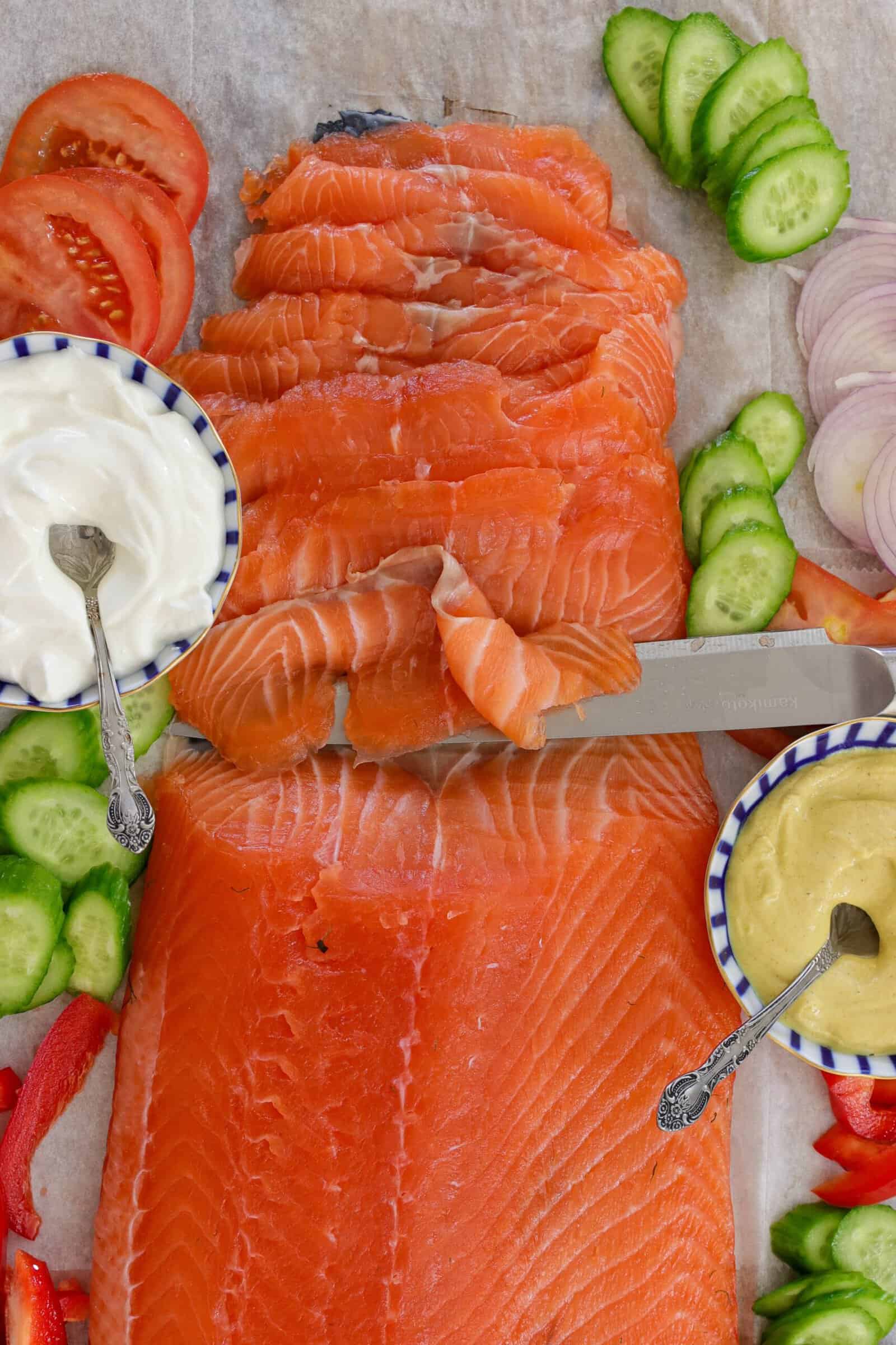 A large slab of cured salmon (homemade gravlax) is surrounded by sliced tomatoes, cucumbers, onions, and bowls of cream sauce and mustard, with a knife slicing the fish.