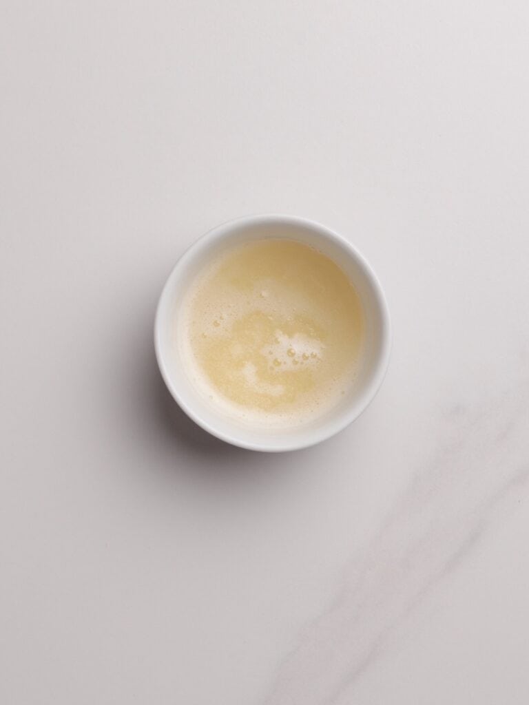 A white bowl filled with a frothy, pale yellow liquid sits on a white marble surface.
