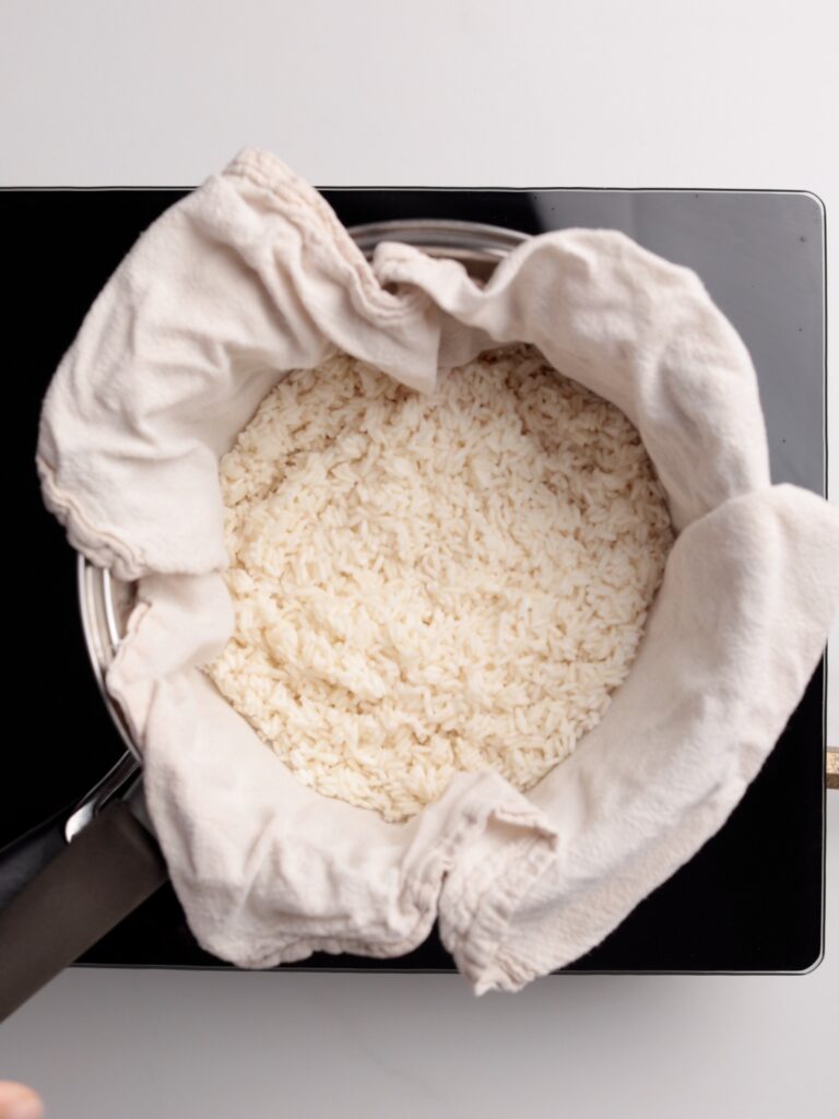 A pot lined with a white cloth is filled with cooked white rice, sitting on a black stovetop against a plain white background.