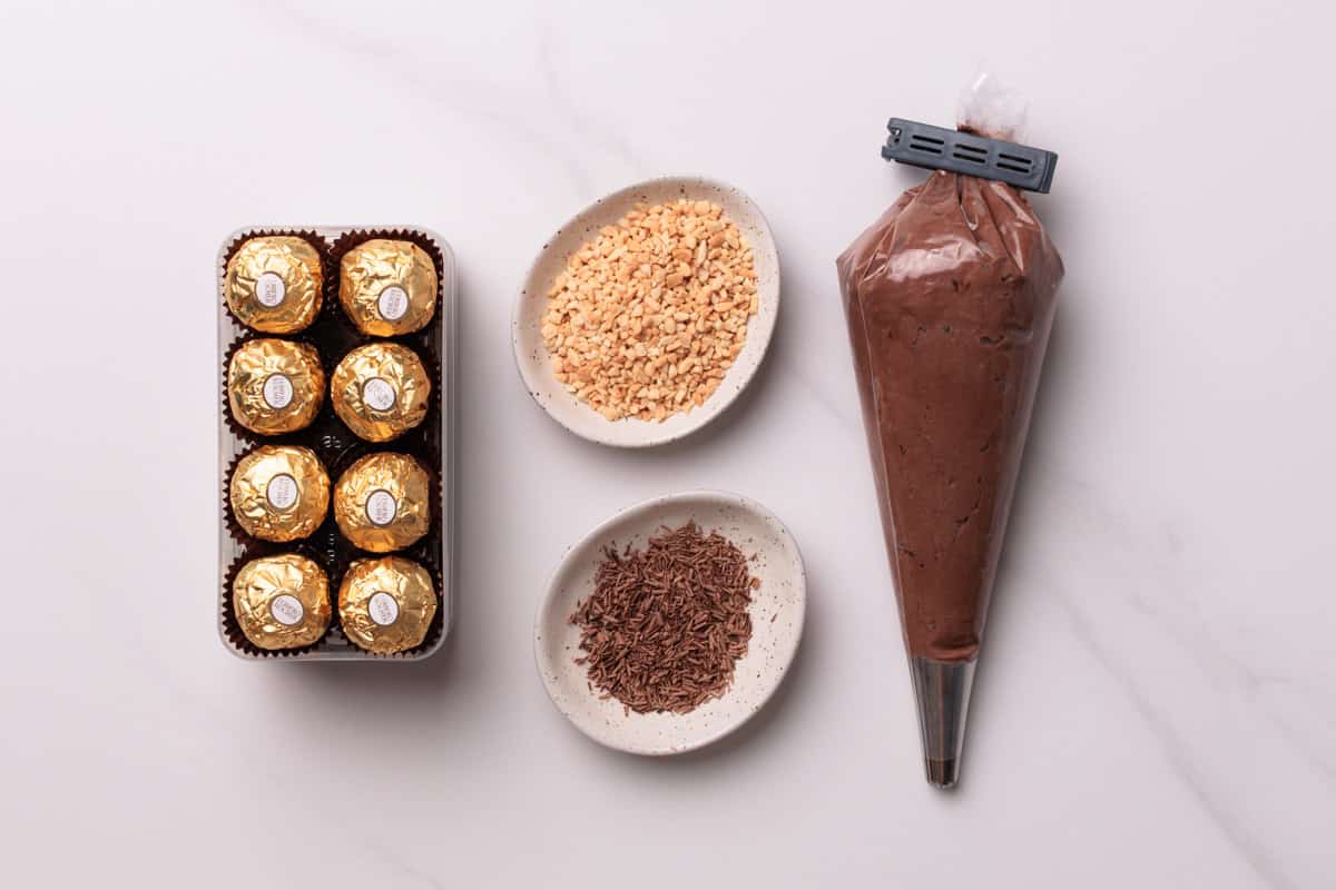 A tray of eight Ferrero Rocher chocolates, a bowl of chopped nuts, a bowl of chocolate sprinkles, and a piping bag filled with chocolate cream arranged on a white surface.