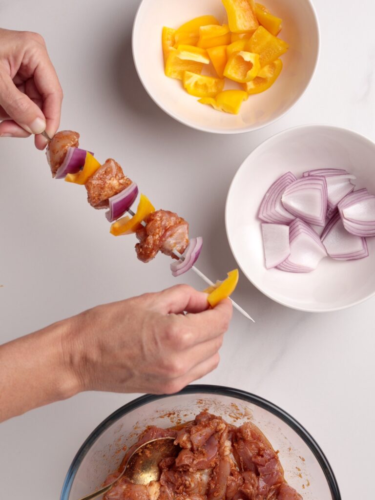 A person is assembling a skewer with marinated chicken, yellow bell pepper pieces, and red onion slices. Nearby bowls hold more yellow bell pepper, red onion, and marinated chicken on a white surface.