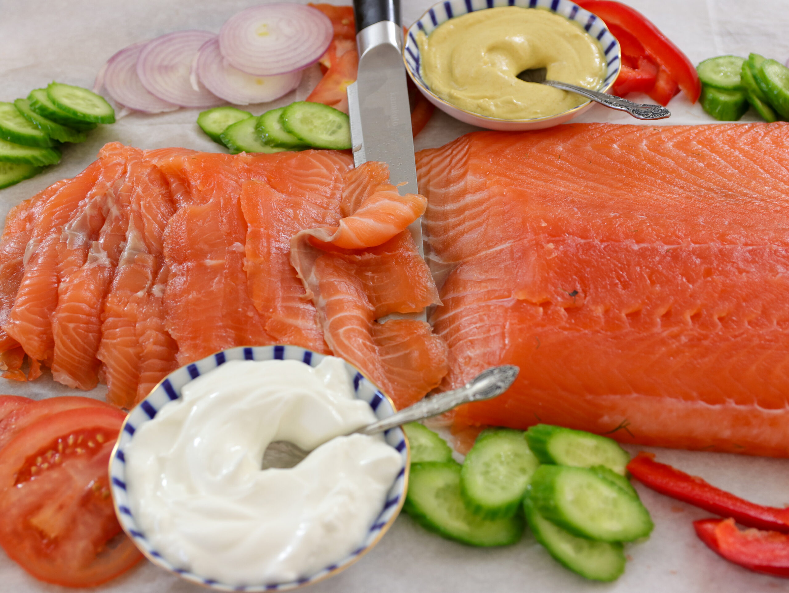 Sliced cured salmon with a knife, surrounded by cucumber, tomato, red bell pepper, onion slices, and two bowls containing cream and mustard sauce.