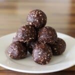 A white plate holds a pyramid stack of chocolate energy bites with dates and visible bits of nuts, set on a wooden table.
