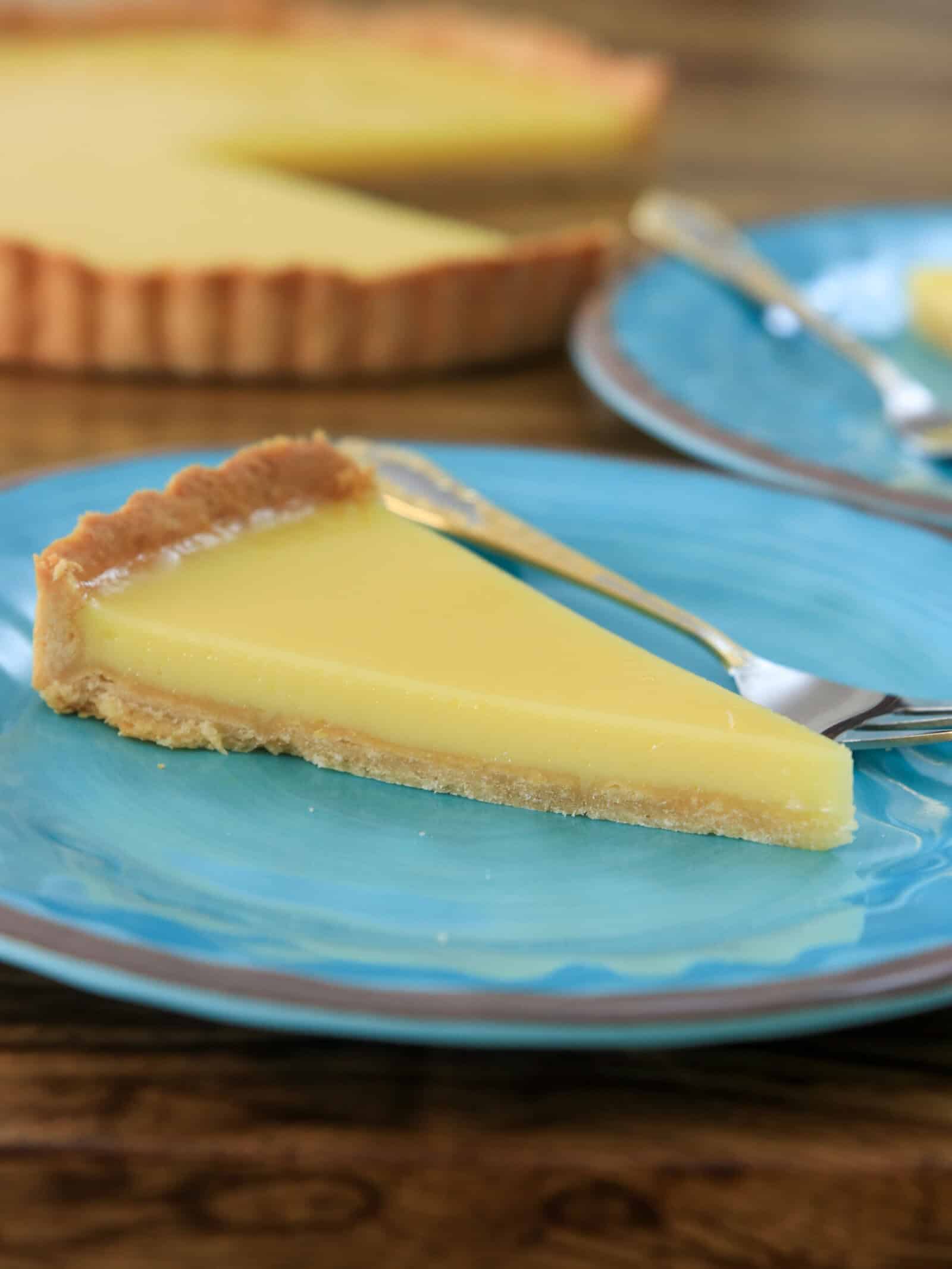 A slice of lemon tart with a golden crust sits on a blue plate with a fork. The rest of the tart is visible in the background on a wooden table.