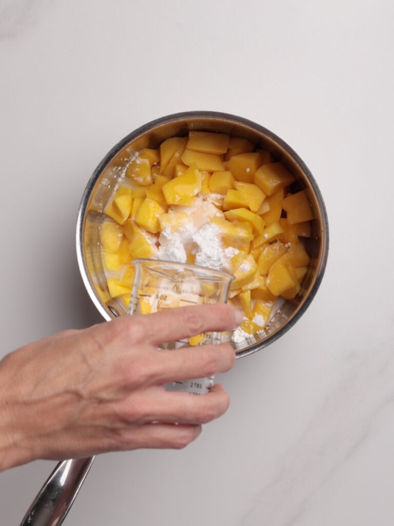 A hand pours liquid from a measuring cup into a pot of diced mango and white powder.