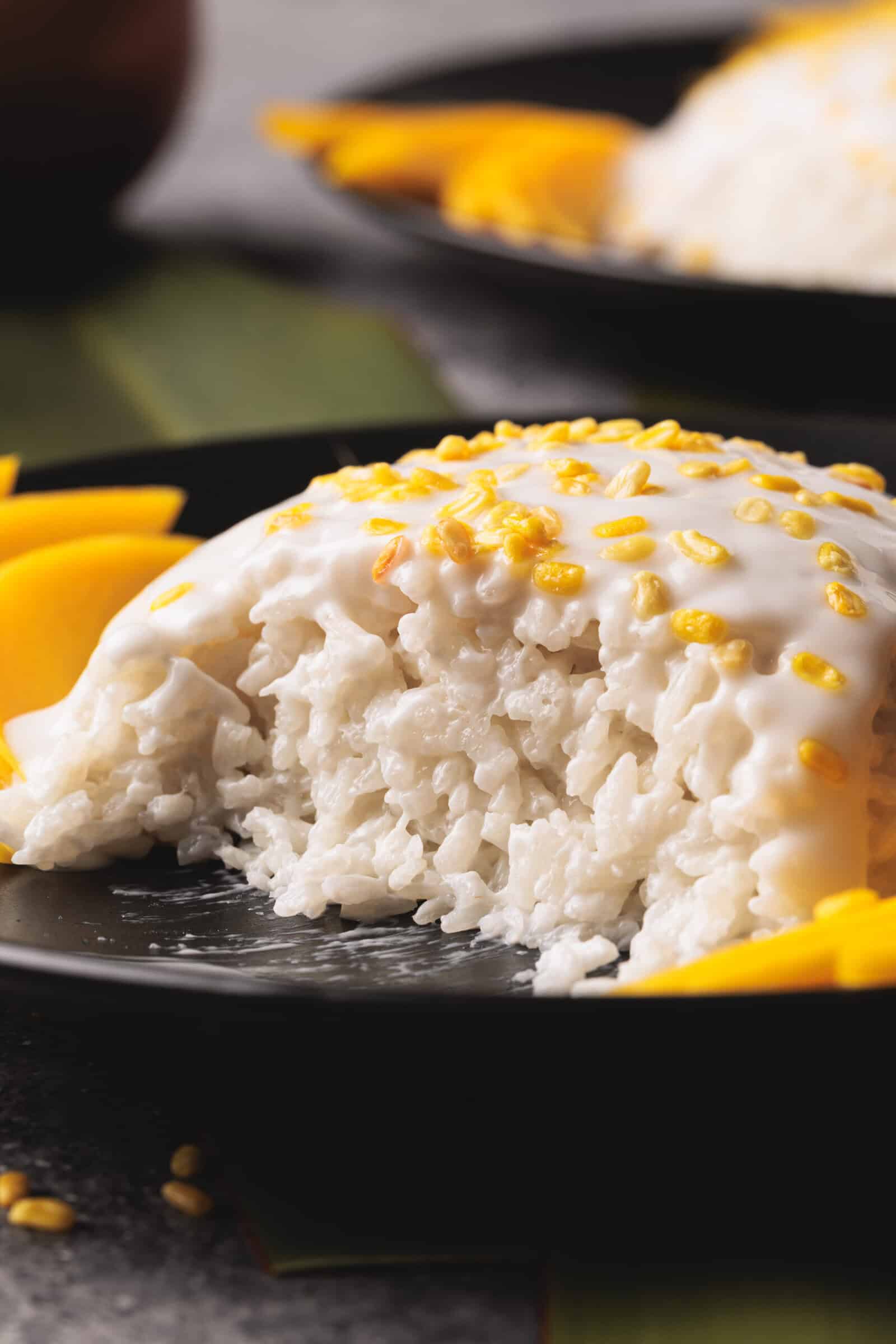 A close-up of mango sticky rice topped with coconut cream and yellow mung beans, with slices of fresh mango on the side, served on a black plate.