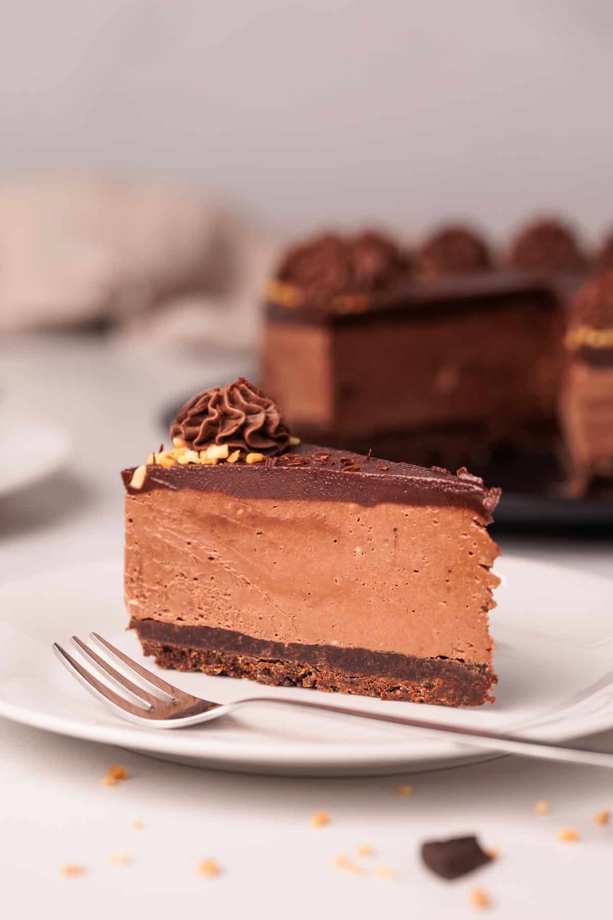 A slice of rich no-bake nutella cheesecake with a chocolate ganache topping and decorative swirl, served on a white plate with a fork. The whole cake is visible in the background.