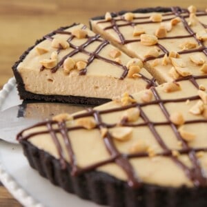 A creamy peanut butter pie with a chocolate cookie crust, topped with a chocolate drizzle and chopped peanuts. One slice is being served from the pie.