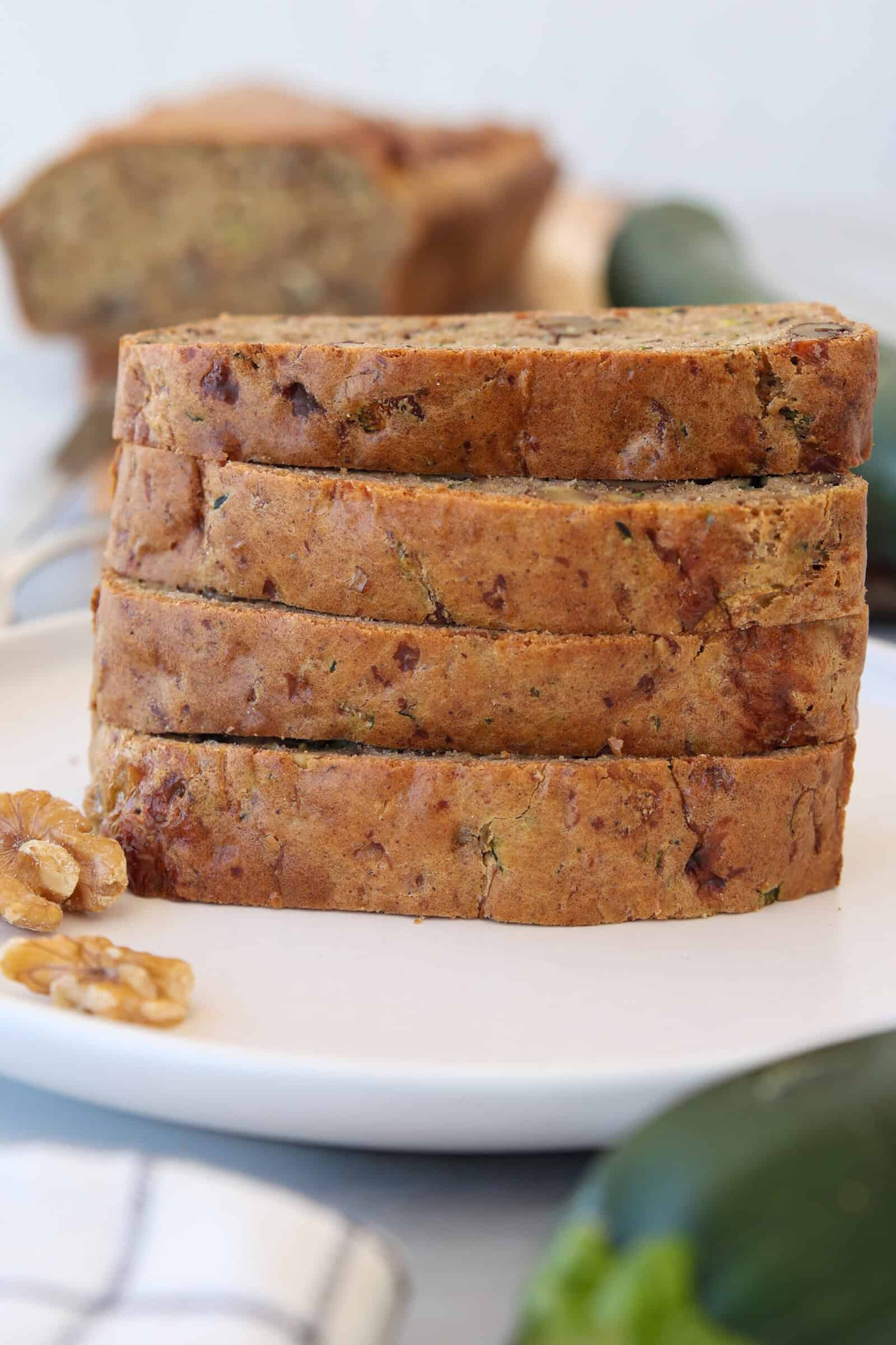 A stack of four slices of zucchini bread sits on a white plate, with walnuts and a whole zucchini nearby. Another loaf is blurred in the background.