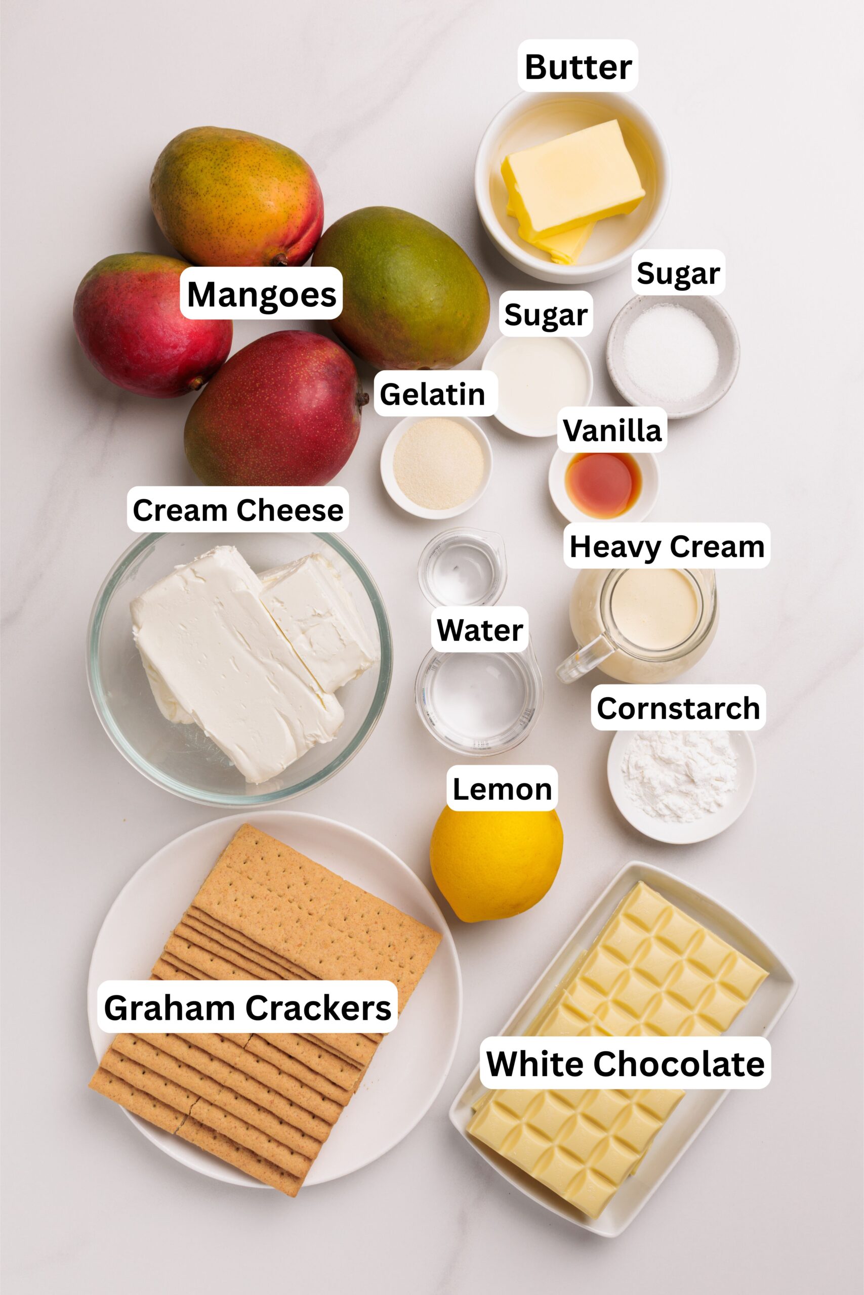 An overhead shot shows ingredients for a dessert, including mangoes, cream cheese, graham crackers, white chocolate, butter, sugar, gelatin, vanilla, heavy cream, water, and a lemon.