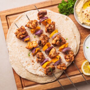 Four skewers with grilled chicken, yellow bell peppers, and red onions are placed on flatbreads, served on a wooden board. Bowls of dip, lemon slices, and fresh herbs are beside them.