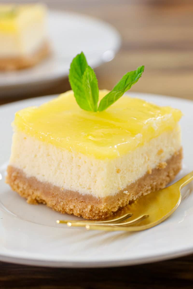 A slice of lemon cheesecake bar with a graham cracker crust, topped with lemon glaze and a sprig of fresh mint, is served on a white plate with a gold fork beside it.