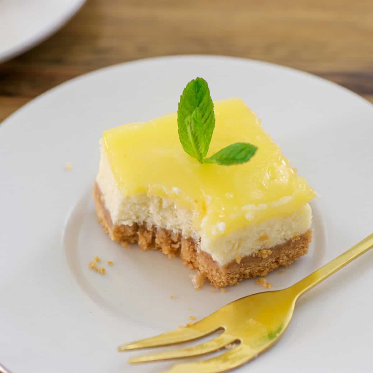 A square piece of lemon cheesecake with a crumbly crust, topped with a yellow lemon layer and a sprig of mint, is served on a white plate with a gold fork.