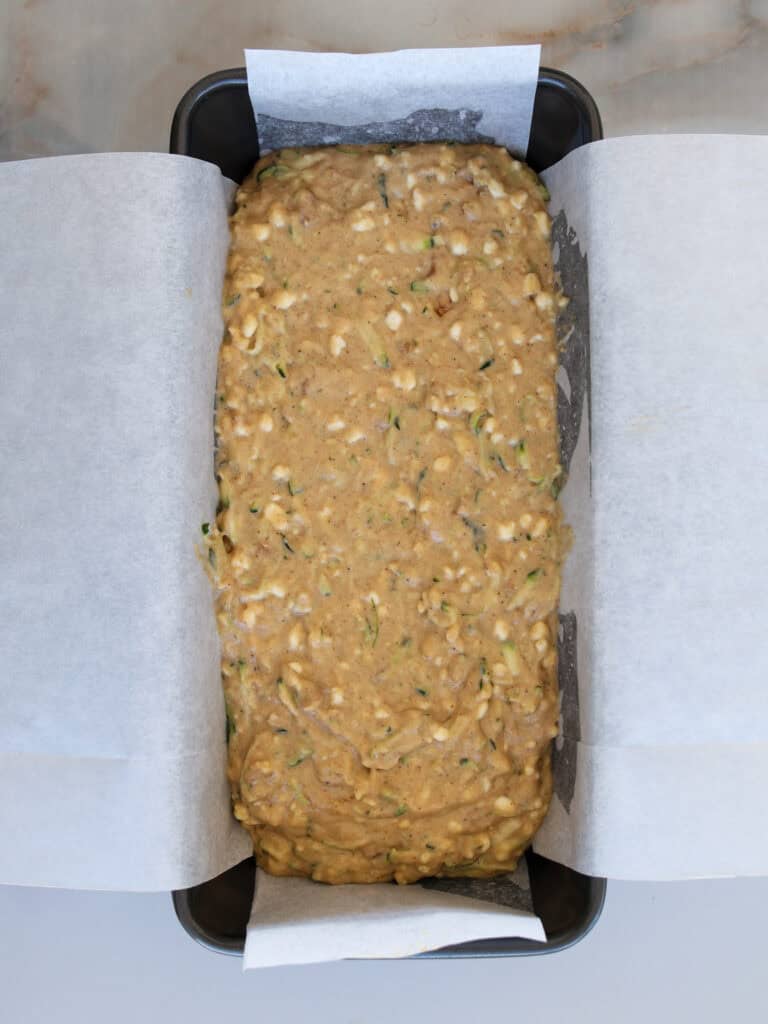 A loaf pan lined with parchment paper is filled with unbaked zucchini bread batter, featuring visible shreds of zucchini and a thick, lumpy texture.