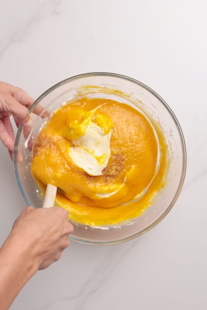 Hands mixing vibrant orange puree with a creamy white mixture in a glass bowl.
