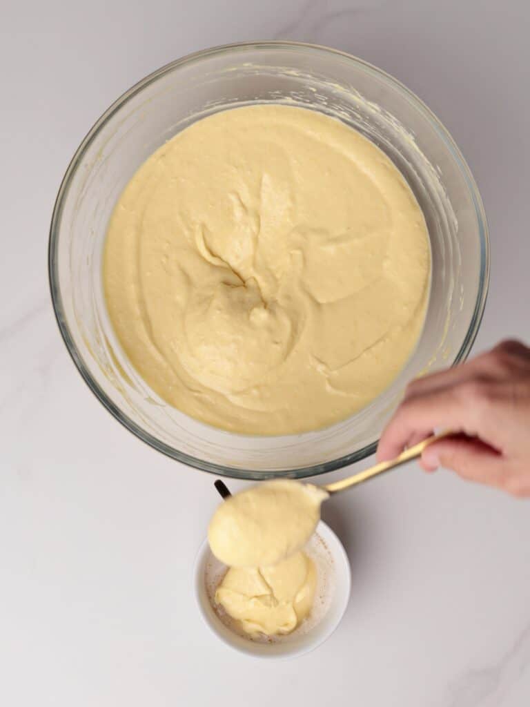 A hand holds a gold spoon to scoop thick, pale yellow batter from a large glass bowl into a small white bowl with frothy liquid.