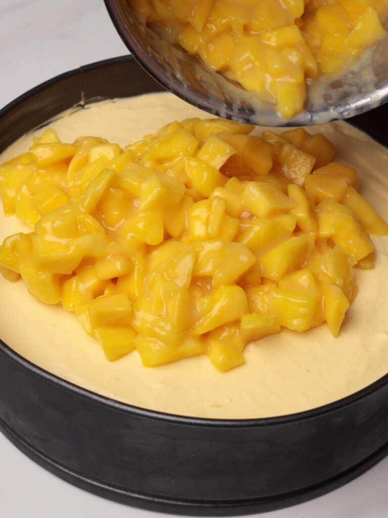 Chopped mangoes being poured into a creamy base in a round baking pan.