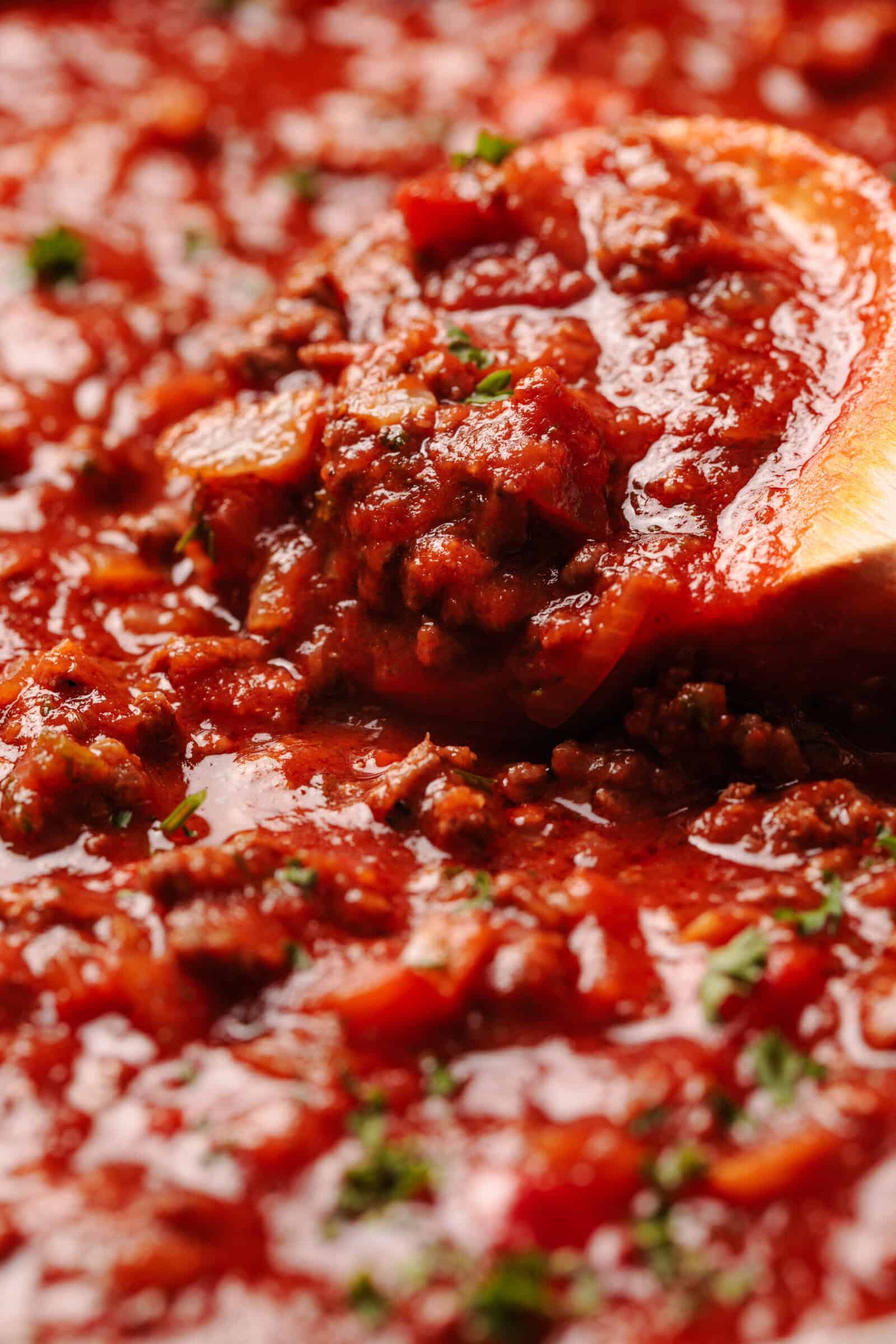 Close-up of a thick, hearty meat sauce for spaghetti being stirred with a wooden spoon, with visible chunks of ground meat and herbs.