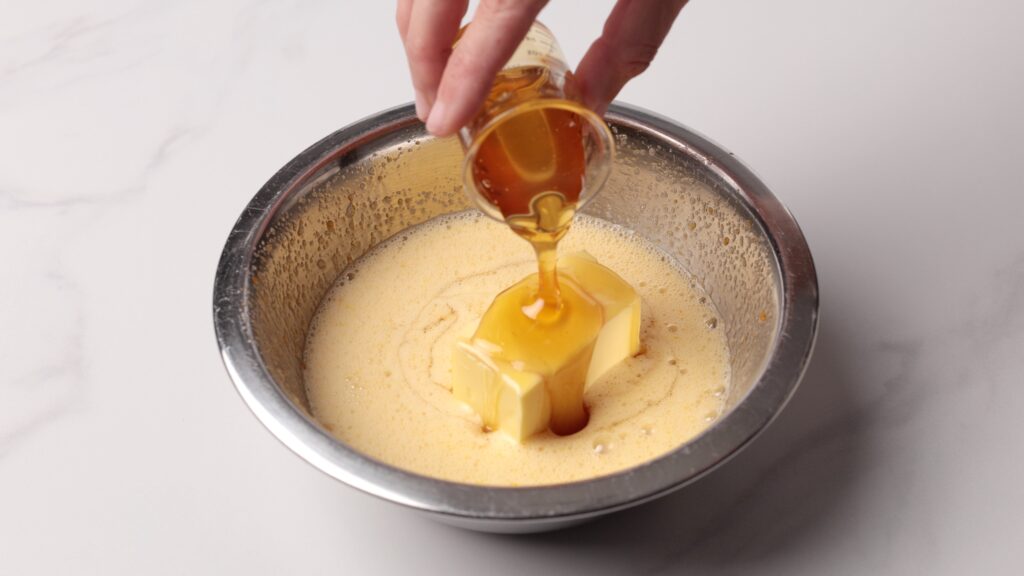 A hand pours honey from a small glass container onto two sticks of butter in a metal bowl containing a creamy, light-colored mixture.