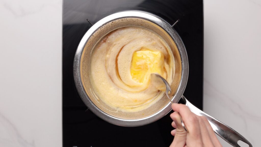 A hand stirs melting butter in a saucepan on a black stovetop, creating a swirl pattern in the foamy liquid.