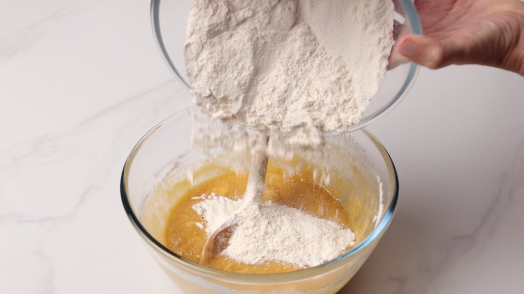 A hand pours flour from a glass bowl into another bowl containing a yellow, wet mixture while stirring with a wooden spoon on a white surface.