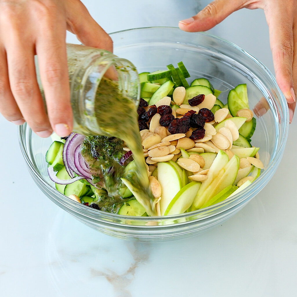 A hand pours green herb dressing from a jar into a glass bowl filled with sliced cucumber, red onion, apple, almonds, and raisins on a white surface.