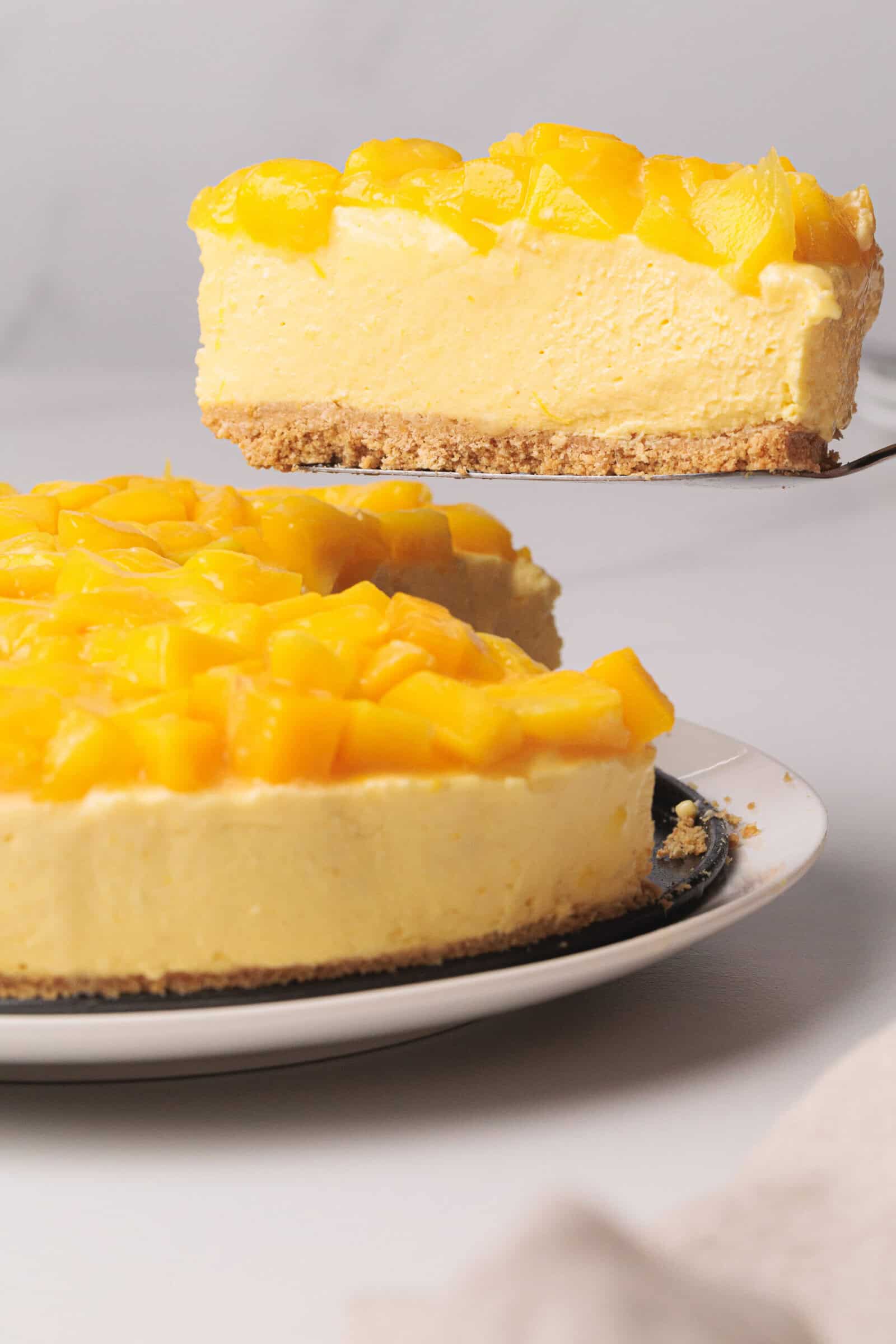 A slice of creamy mango cheesecake is lifted from the whole cake, both topped with fresh mango chunks.
