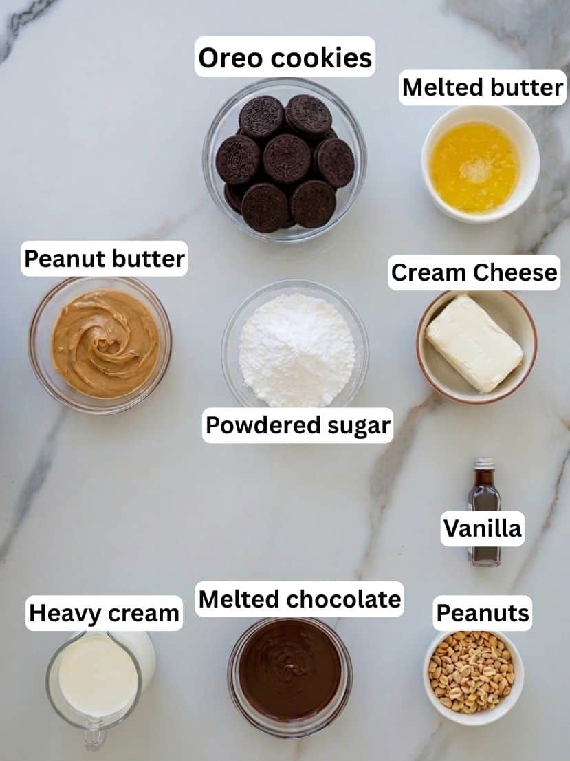Top-down view of labeled ingredients for a dessert on a marble surface, including Oreo cookies, melted butter, peanut butter, powdered sugar, cream cheese, vanilla, heavy cream, melted chocolate, and peanuts.