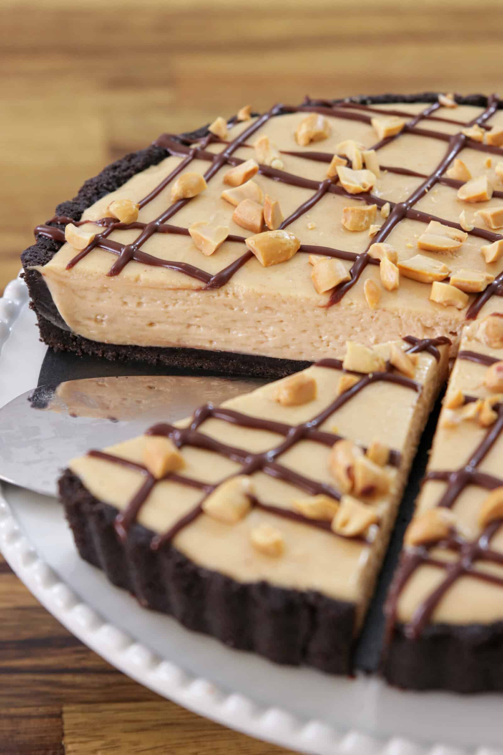 A creamy peanut butter pie with a chocolate cookie crust, topped with a chocolate drizzle and chopped peanuts, sits on a white plate with one slice being served.