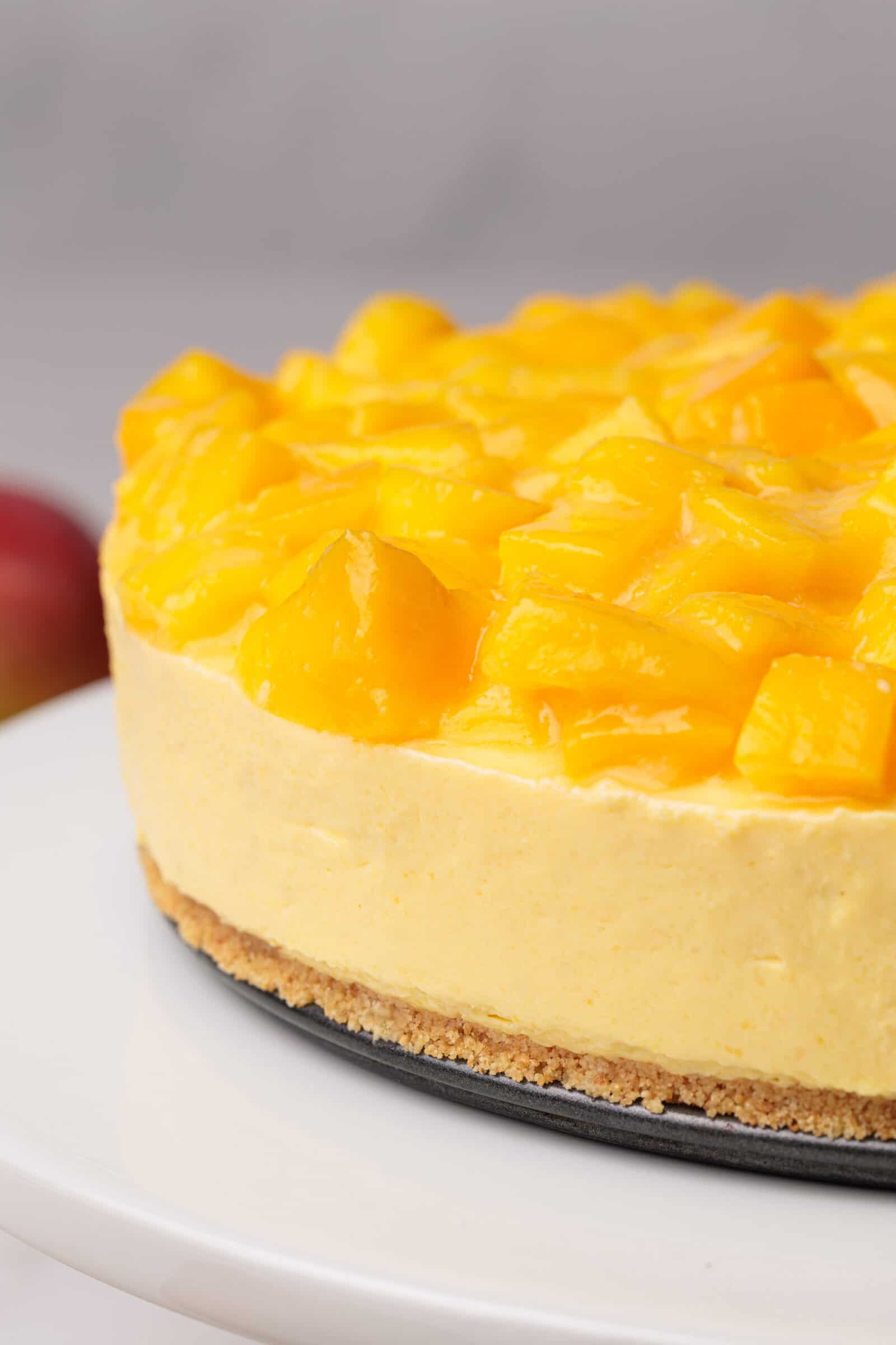 A whole n0-bake mango cheesecake topped with fresh diced mango chunks sits on a white cake stand.