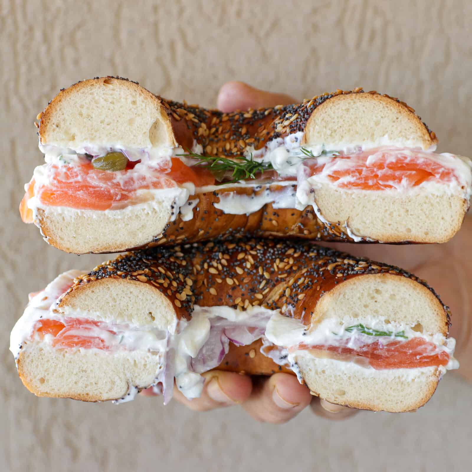 A hand holds two halves of a seeded bagel sandwich filled with cream cheese, cured salmon, capers, red onion, and fresh herbs, with the fillings visible against a neutral background.