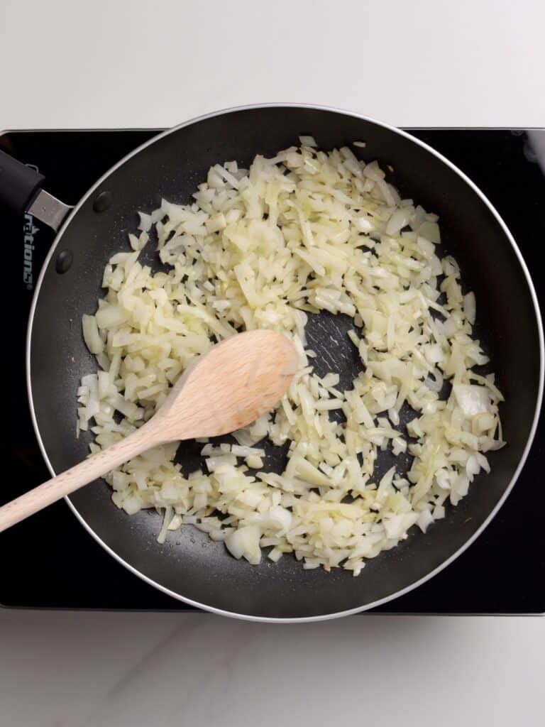 Chopped onions being sautéed in a black frying pan on a stovetop, stirred with a wooden spoon.
