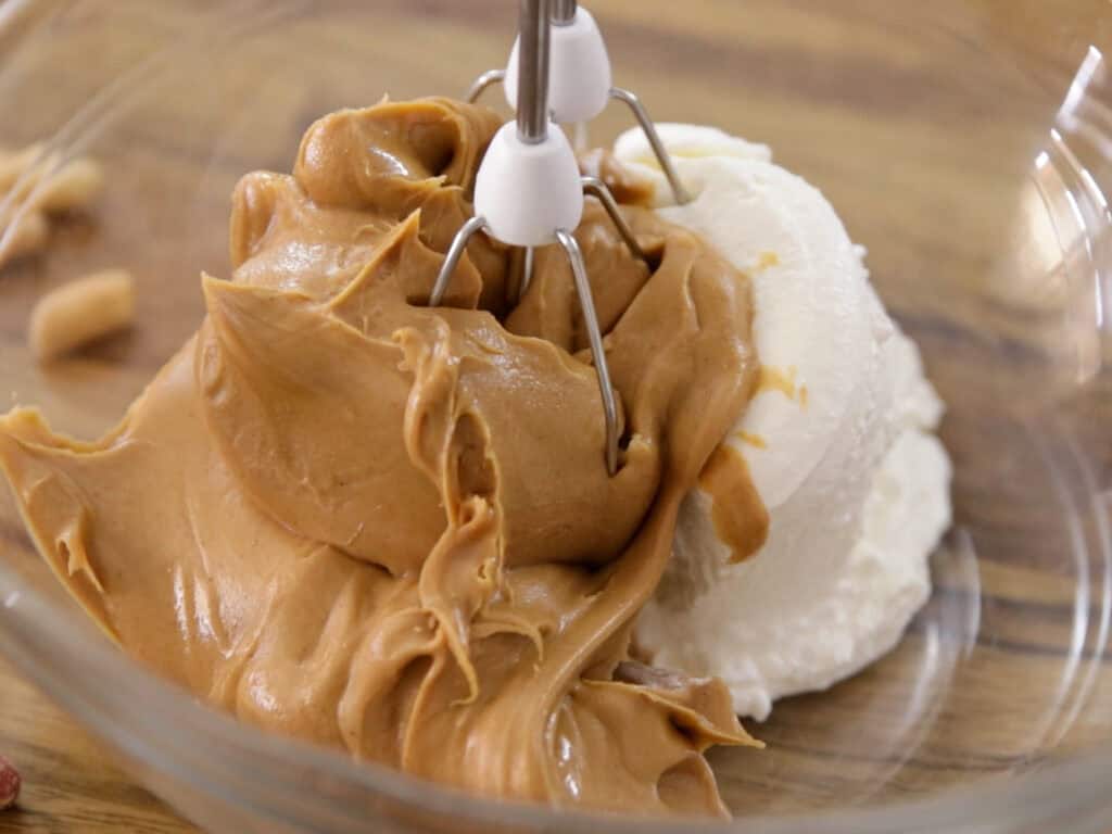 A close-up of an electric mixer in a glass bowl containing creamy peanut butter and a scoop of white cream cheese, ready to be blended together on a wooden surface.