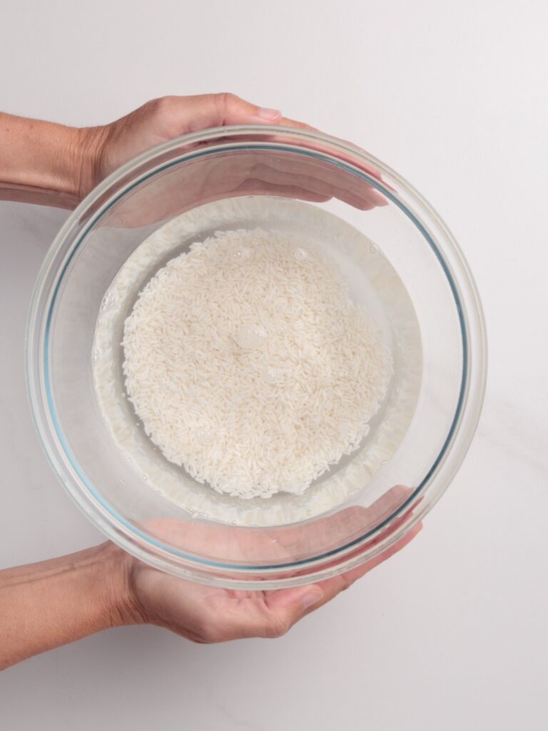 A pair of hands holds a clear glass bowl filled with water and uncooked white rice, seen from above on a light-colored surface.