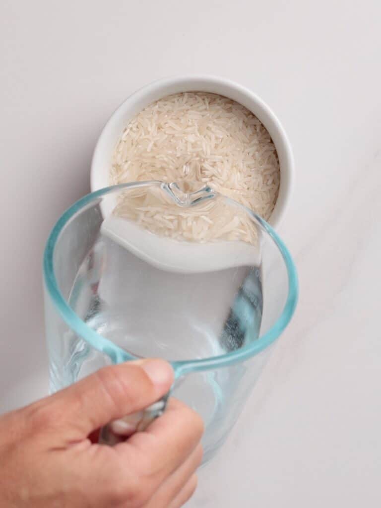 A hand pours water from a clear glass pitcher into a white bowl filled with uncooked white rice on a white surface.