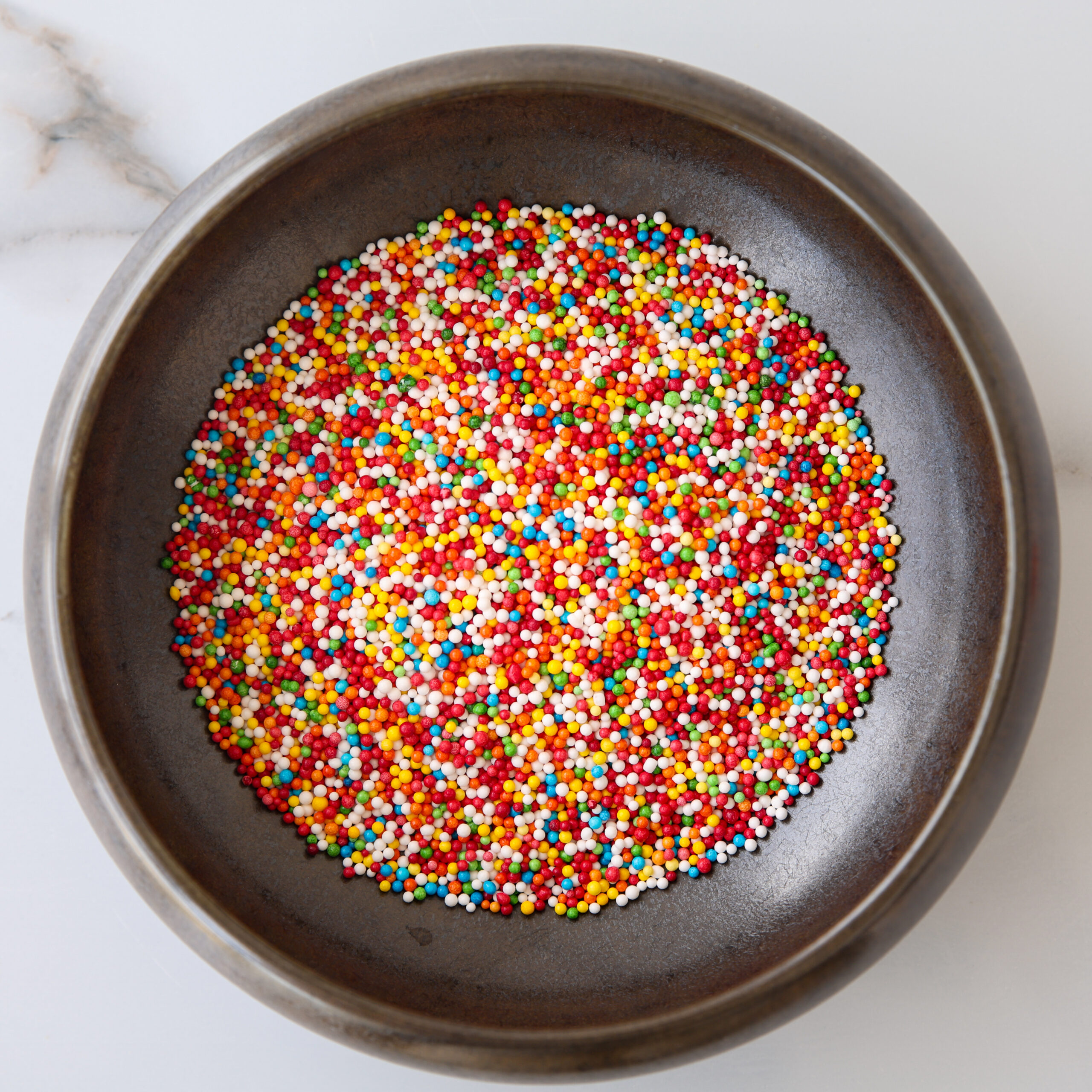 A dark bowl filled with colorful round sprinkles in shades of red, yellow, green, blue, orange, and white, sitting on a light marble surface.