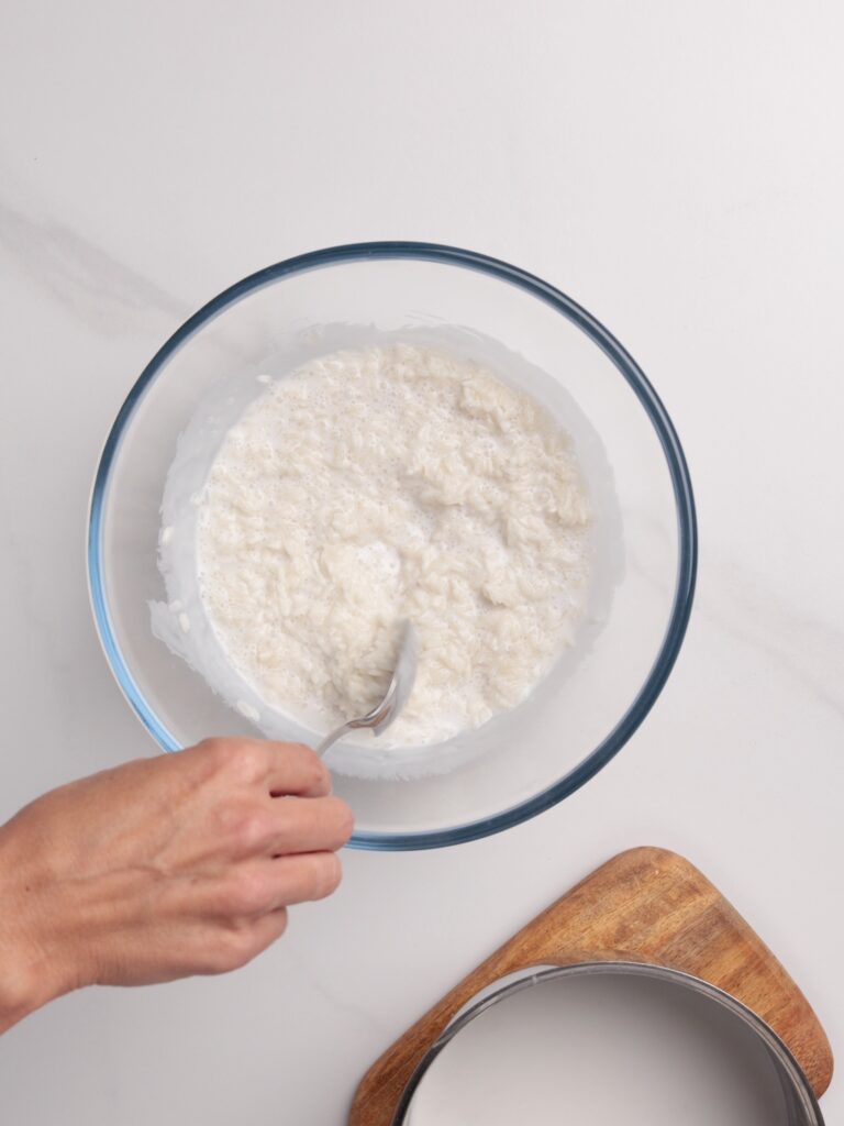 A hand holding a spoon stirs a white, creamy mixture in a glass bowl on a white surface. A wooden board with a metal ring and a bowl are nearby.
