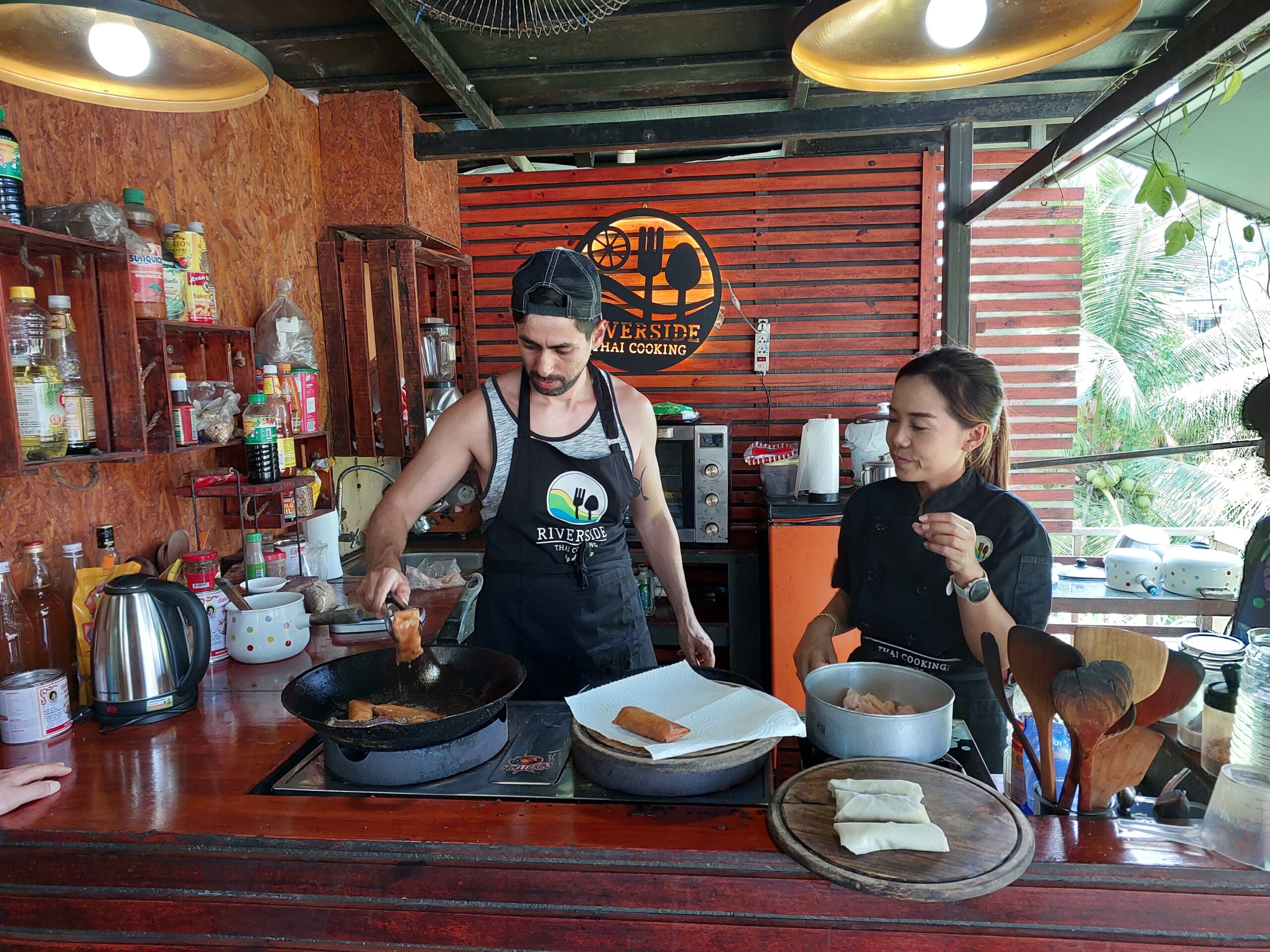 Two people in a rustic kitchen prepare food together. David is frying something in a pan, while the woman (Thai cooking master) watches. The kitchen features wooden shelves, jars, utensils, and a "Riverside Thai Cooking" sign.