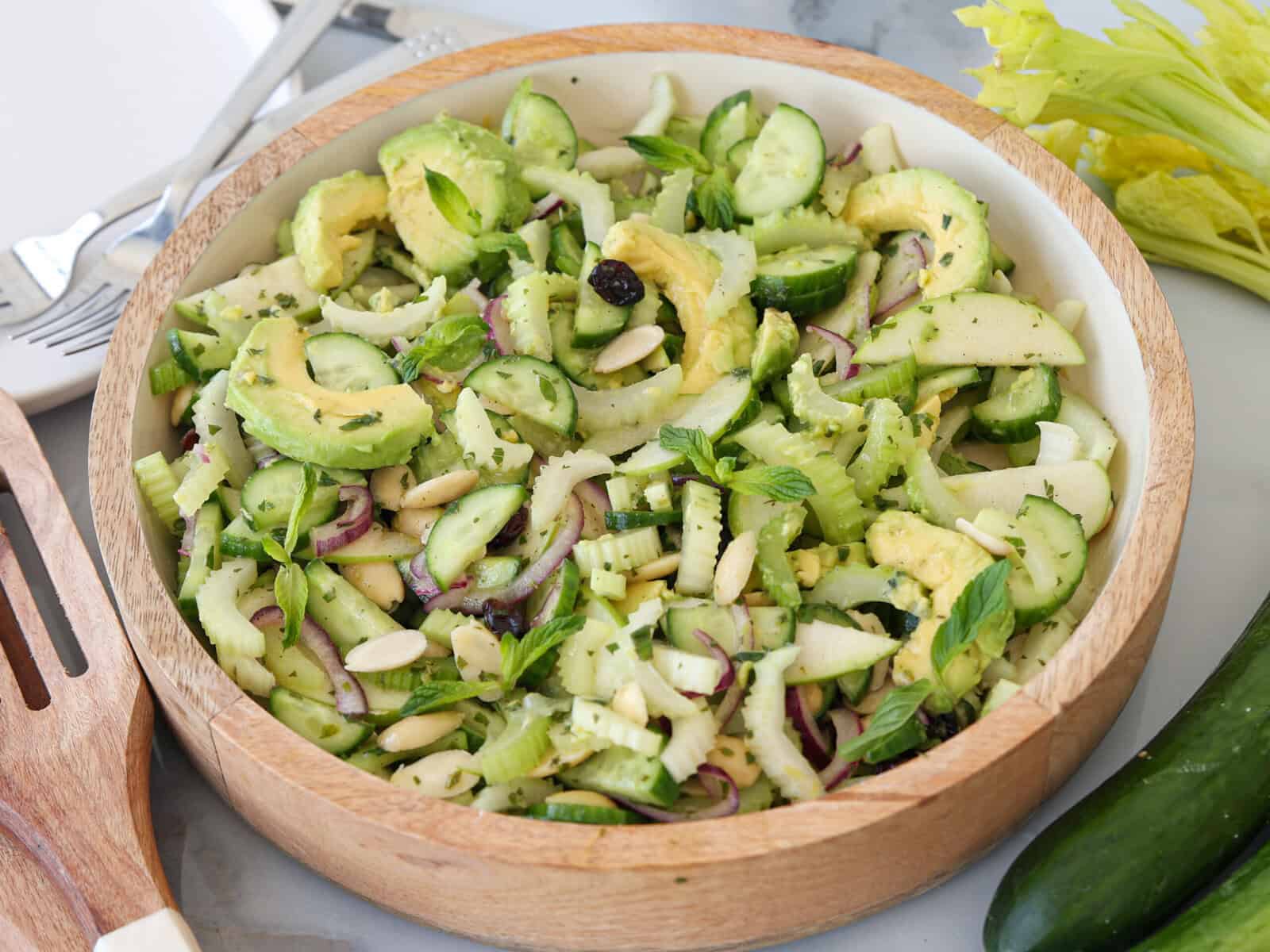 A fresh salad in a wooden bowl, featuring sliced cucumber, avocado, celery, apple, red onion, mint leaves, nuts, and herbs. A wooden serving utensil and celery stalk are nearby on a light surface.