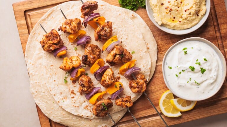 Three baked chicken and vegetable skewers on flatbreads, served on a wooden board with bowls of white yogurt sauce and hummus, garnished with herbs and lemon slices on the side.