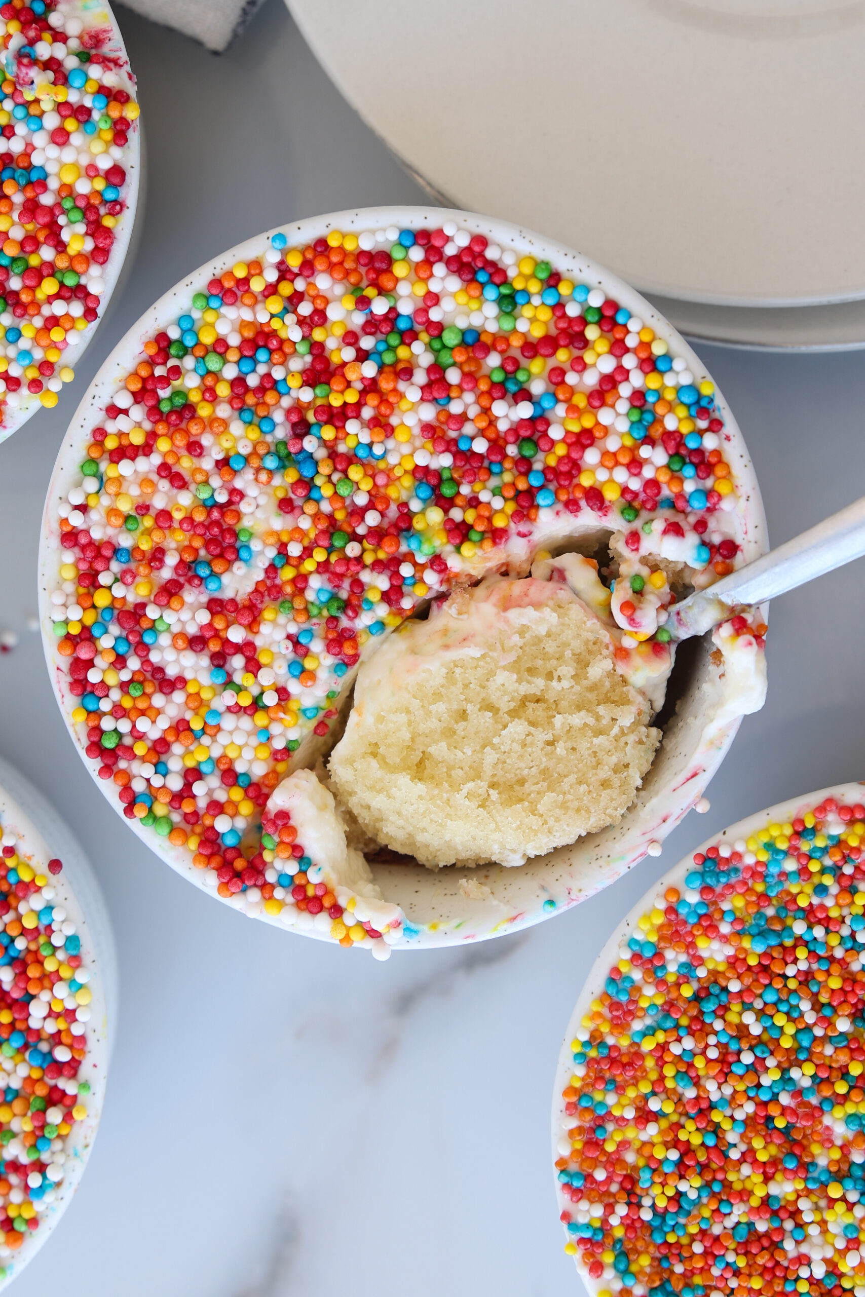 A viral homemade dot cake filled with vanilla cake topped with white frosting and colorful round sprinkles. A spoon has scooped out a portion, revealing the soft cake inside. The mug sits on a light surface near plates.