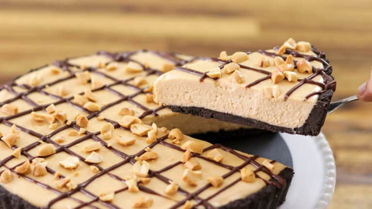 A creamy peanut butter pie with a chocolate cookie crust, topped with chopped peanuts and chocolate drizzle, with one slice being lifted from the pie.