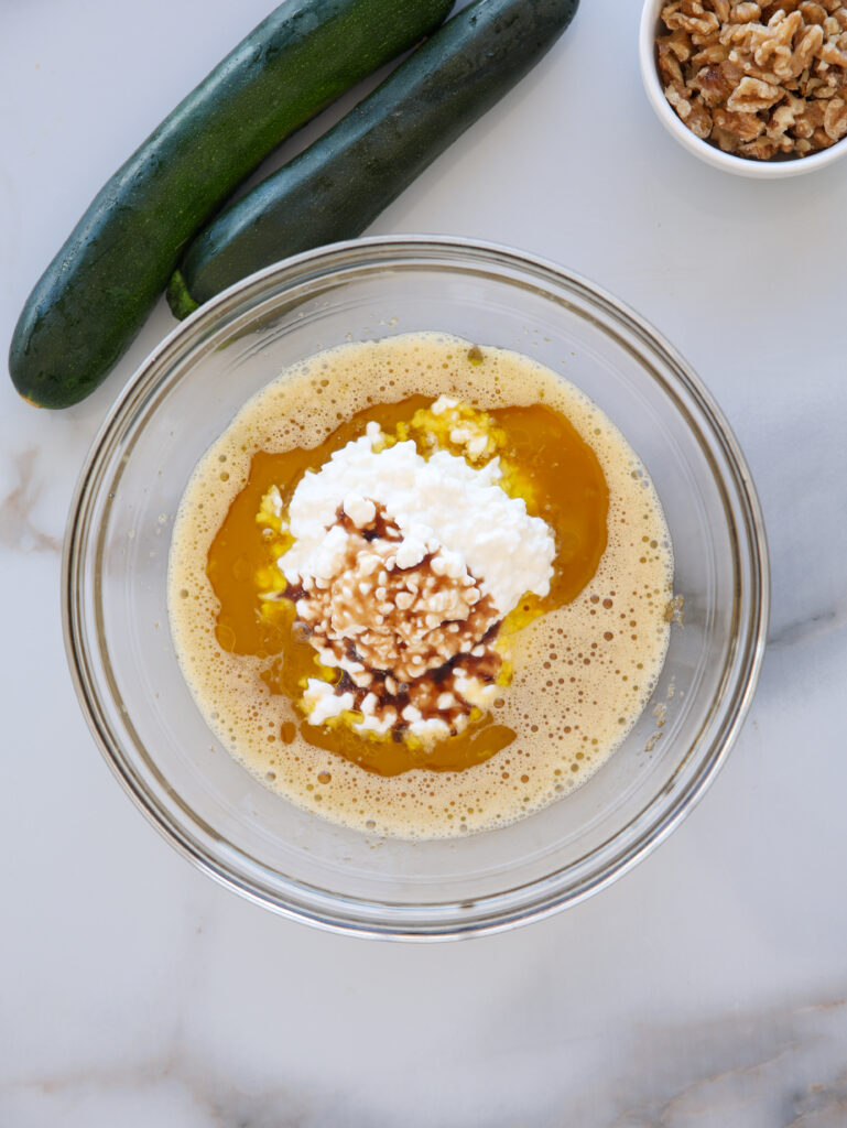 A glass bowl contains a mixture of eggs, cottage cheese, chopped nuts, and vanilla extract. Next to the bowl are two zucchinis and a small dish of walnuts on a white surface.