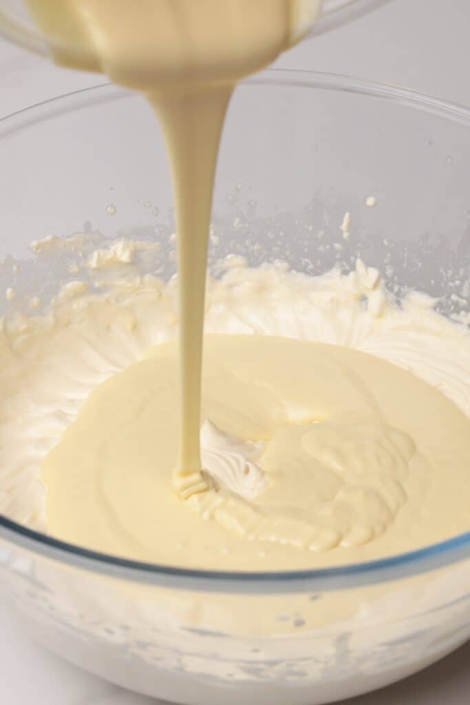 Creamy yellow liquid being poured from a pitcher into a glass bowl filled with whipped cream.