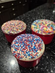Three red cupcake liners filled with cupcakes topped with white frosting and colorful sprinkles—one with red, white, and blue sprinkles, and two with mixed rainbow sprinkles—on a dark granite countertop.