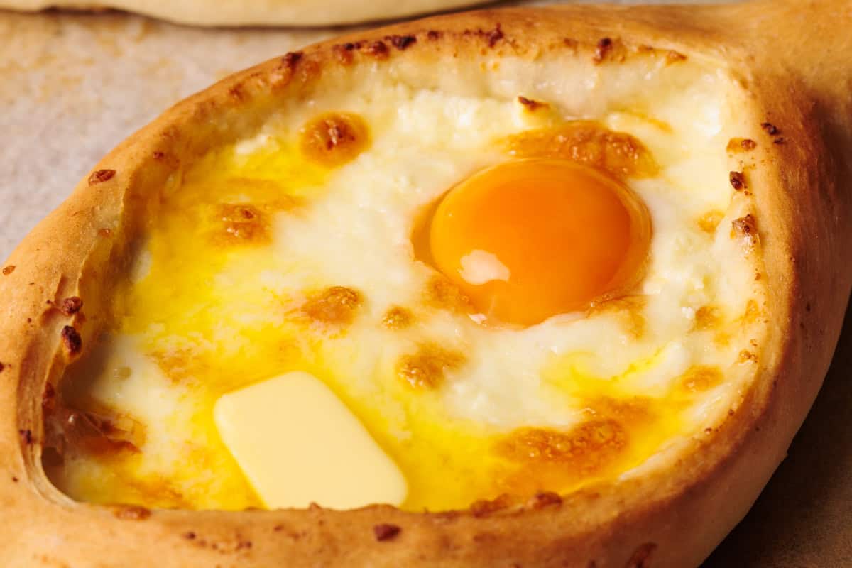 A close-up of khachapuri, a Georgian bread boat filled with melted cheese, a sunny egg yolk, and a pat of butter on top.