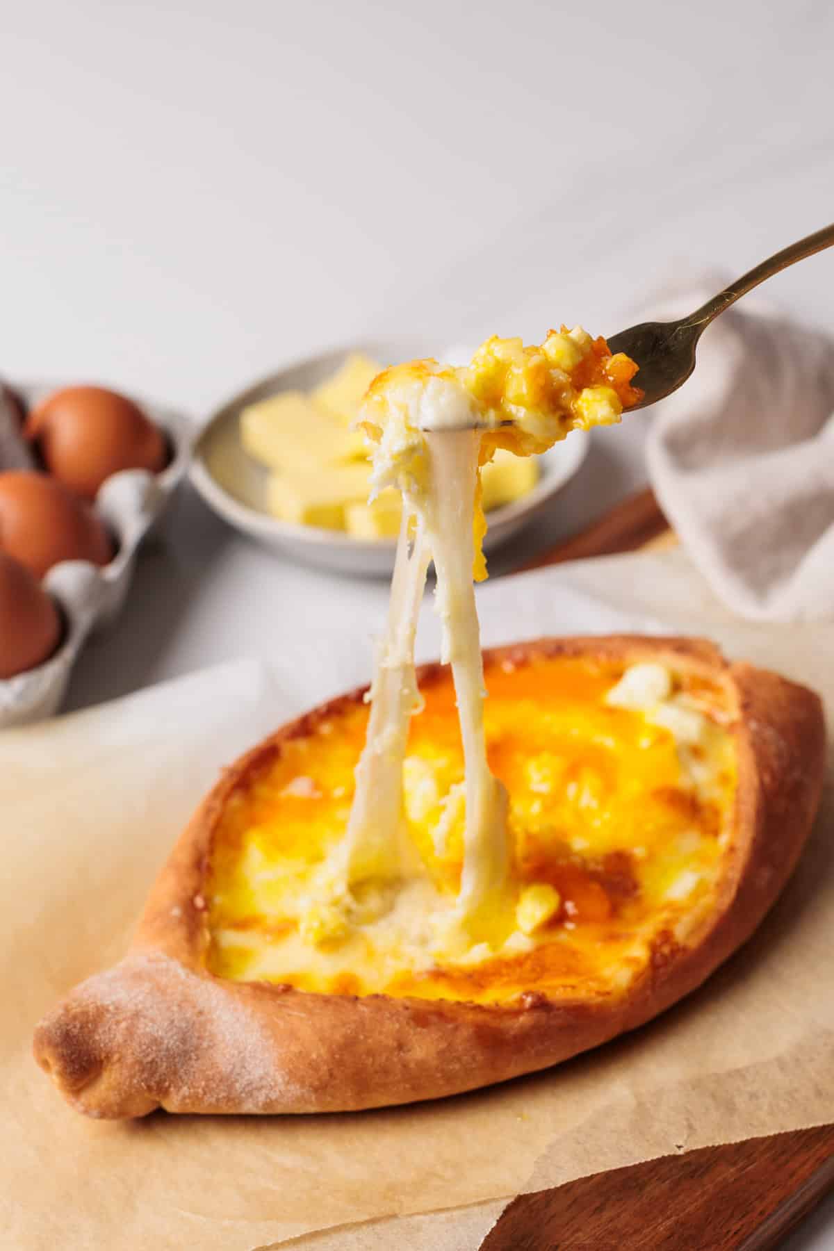 A fork lifts gooey melted cheese and egg from a baked khachapuri bread, with eggs and butter on a plate in the background.
