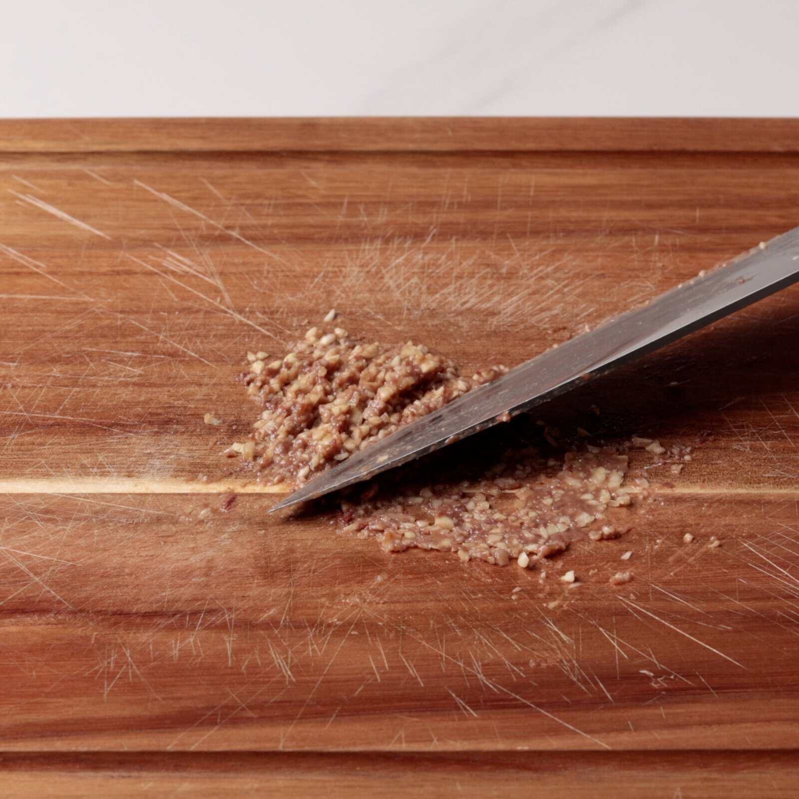 A close-up of a knife mincing anchovies on a scratched wooden cutting board.