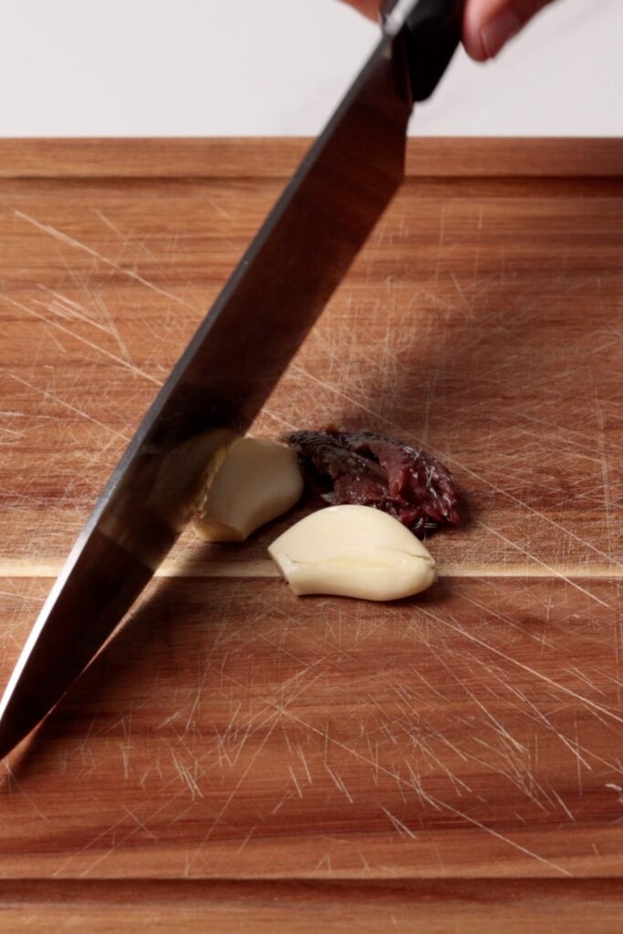 A large knife presses down on garlic cloves and what appears to be an anchovy fillet on a scratched wooden cutting board.
